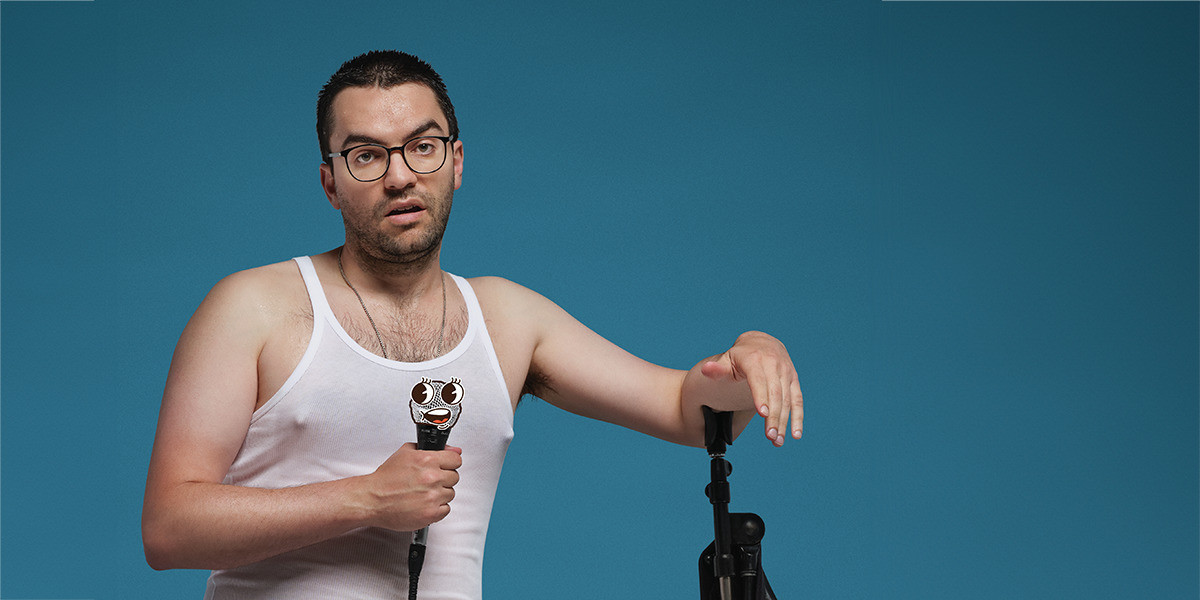 Andrew Portelli - Real Talk - Man with glasses holding a microphone and resting his other arm on the mic stand