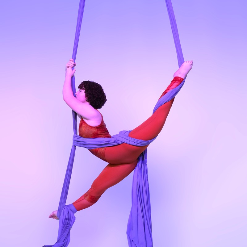 Young woman, in orange costume, wrapped in purple material doing splits