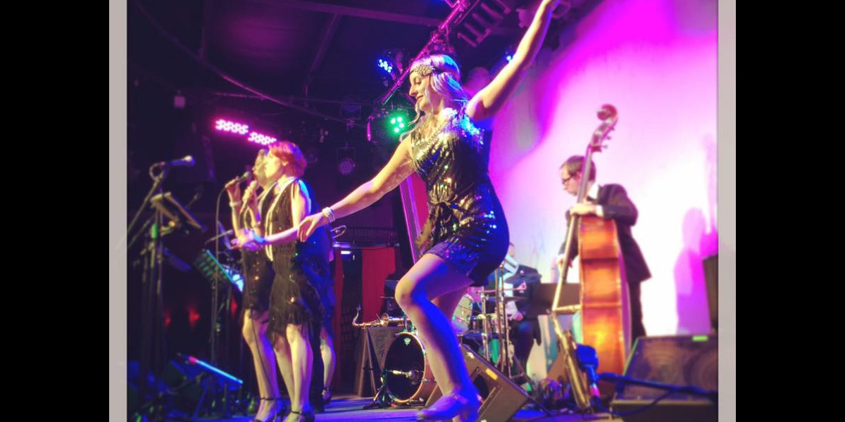 a colourful image of a jazz band on stage. in the foreground is a tap dancer, she has her hands in the air and is mid step, her long blonde hair flows around her.