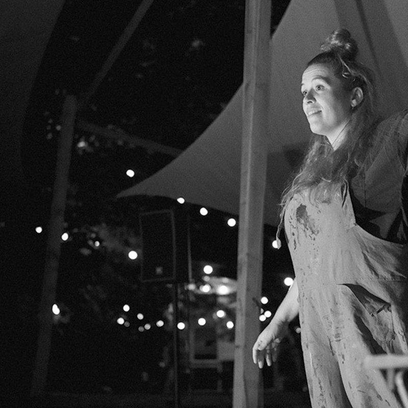 Casey Jay Andrews stands on an outdoor stage surrounded by festoon lighting. She is pointing with her arm outstretched.