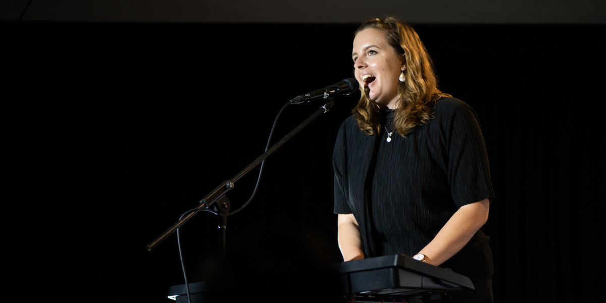 A woman wearing a black t-shirt plays an electric keyboard facing out towards an audience on a stage. She is singing into a microphone on a stand with her mouth wide open and a small smile on her face. There is a black curtain behind her.