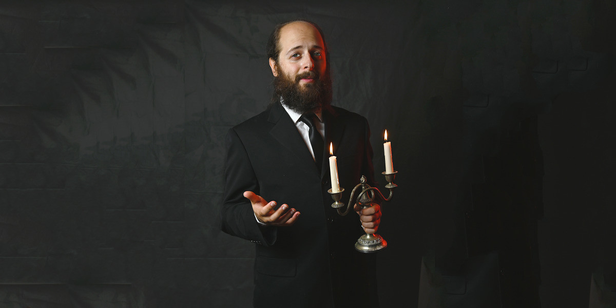 Ben Volchok stands in a black suit and tie, holding a candelabra with two lit candles, and one hand outstretched inviting you to join him.