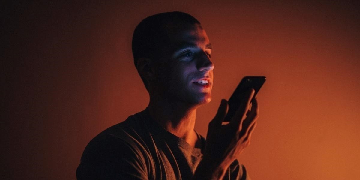 An actor, wearing a dark t shirt and bathed in orange light, holds their smart phone in front of them - talking into it.