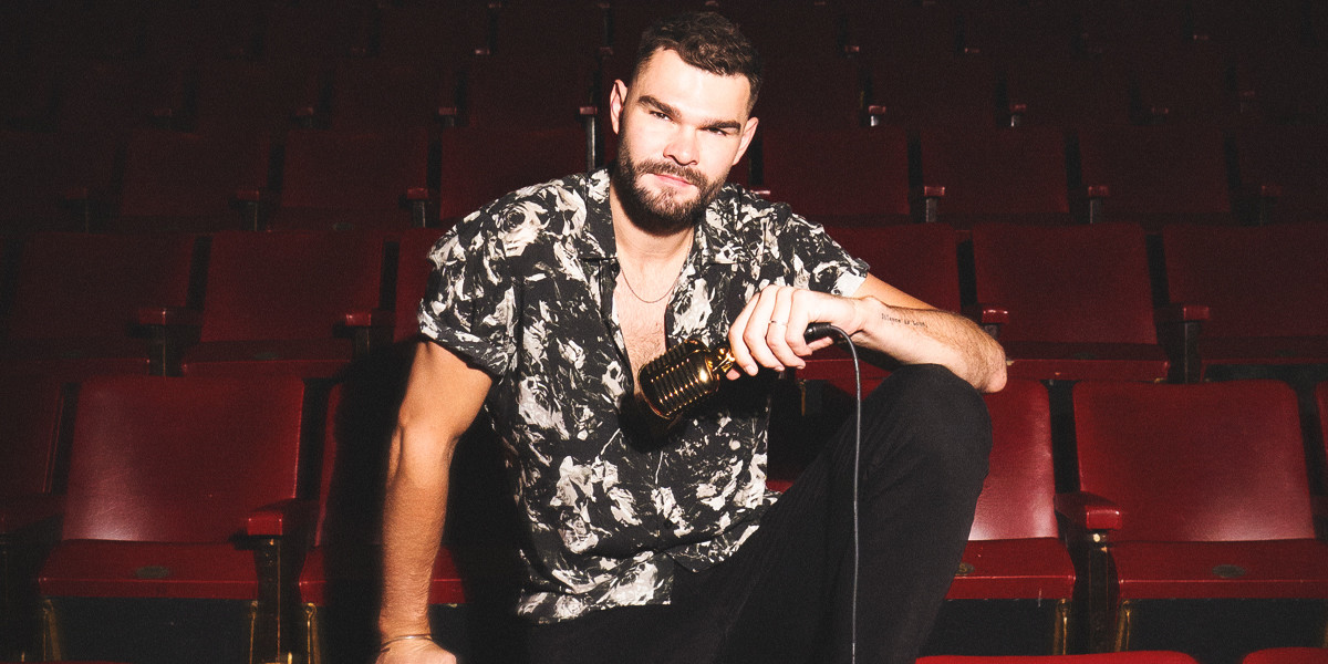 A man sits in a theatre with red vinyl seats loosely holding a 1920s silver microphone as if ready to start singing.