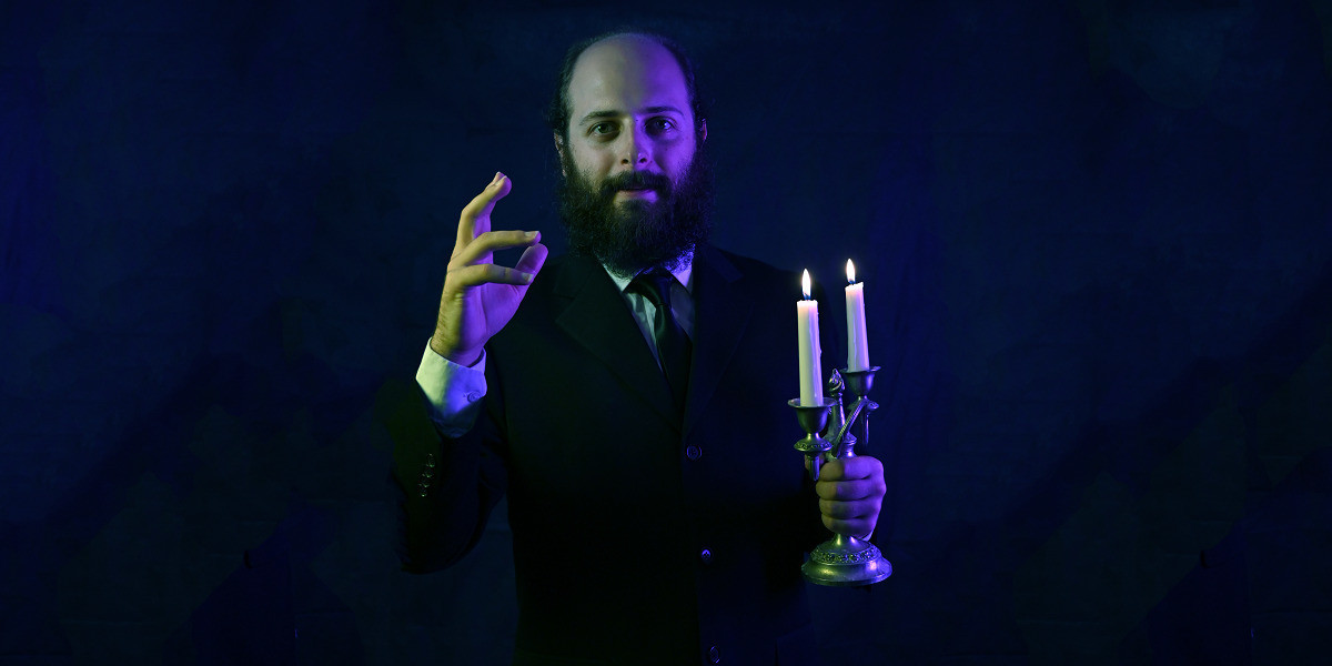 Ben Volchok stands in a black suit and tie, holding a candelabra with two lit candles, and one hand in a ceremonial gesture. The background is purple and green.