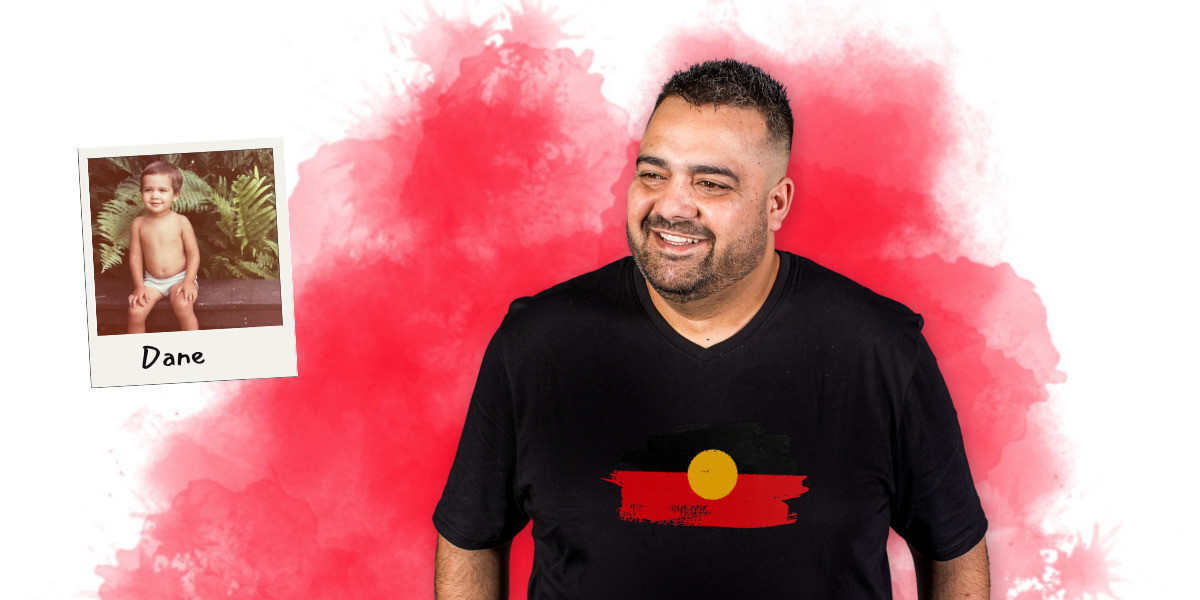 Aboriginal comedian Dane Simpson wears a black t-shirt with Aboriginal flag on the front. He looks off to one side toward a polaroid photo of himself as a young child in front of some ferns.
