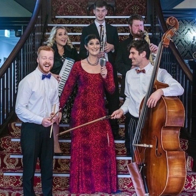 6 people, 2 women and 4 men, standing on stairs holding their instruments- double bass, trumpet, saxophone, drum sticks and key board, lead female singer holding old fashioned microphone. Men dressed in black suits and tie, women formal dress's. Everyone is smiling and having fun looking a bit cheesy !