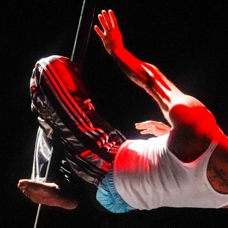 A man wearing a white singlet and baggy pants is suspending themselves from a circus pole