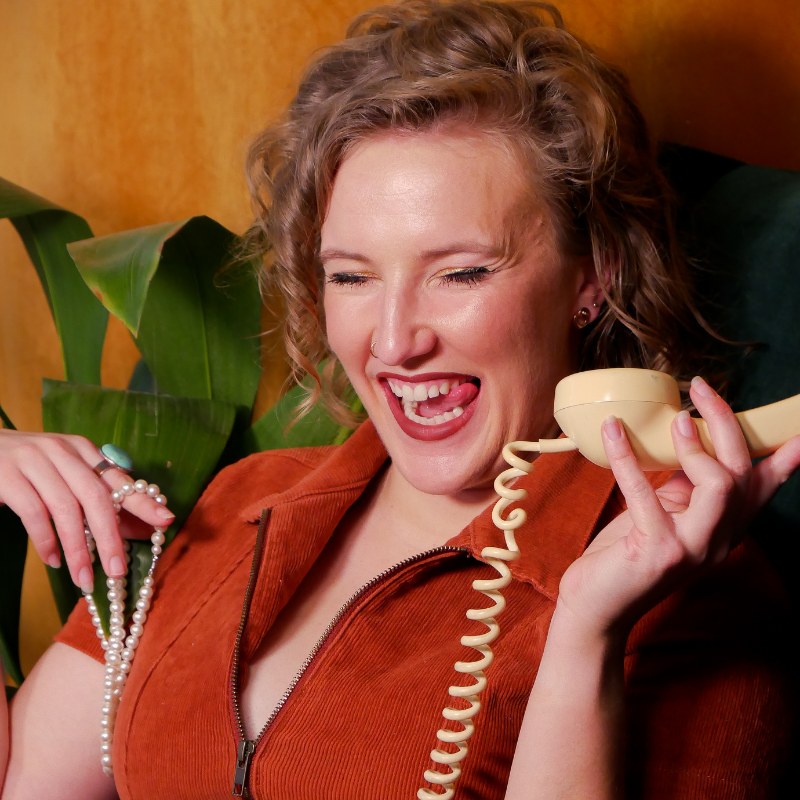 Mim, a white woman with curly, dark blonde hair is wearing an orange-coloured jumpsuit and sitting in a dark green armchair in front of a mustard-coloured background. She is holding the receiver of a retro telephone to her ear, and is laughing with her eyes shut. Her other hand is sporting a large turquoise ring and holding a string of pearls. Behind her are the leaves of an indoor plant.