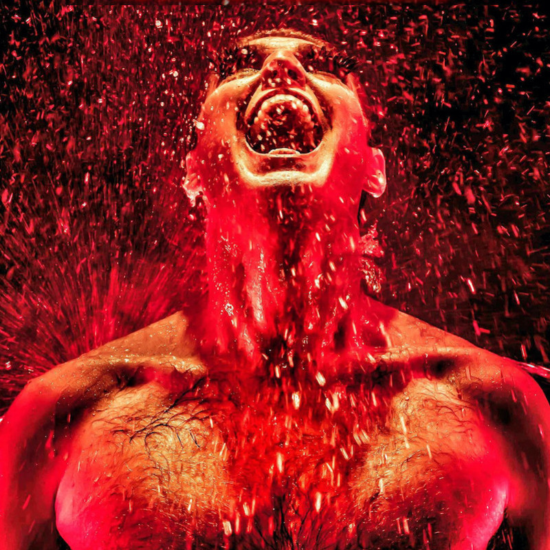 Reuben Kaye, man in drag makeup is looking up mouth wide open, with water splashing down on him, under a red light