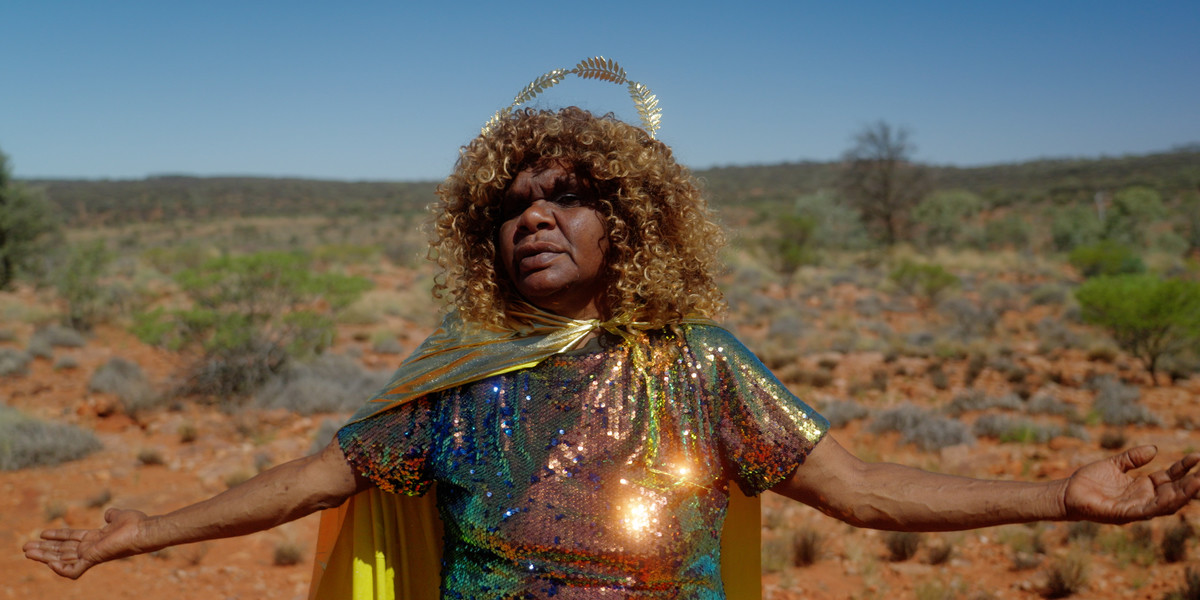 Between the Details: Video Art from the ACMI Collection - A photographic portait of Kaylene Whiskey in a desert landscape, with her arms open, she appears basking in the sun which reflects bright light off her sequined outfit and golden cape.
