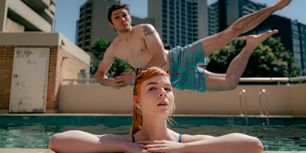 A young man jumps into a swimming pool behind a young woman leaning on the edge of the pool.