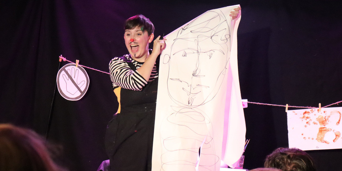 An artist, in black overalls with a black and white striped shirt, red paint splodge nose and drawn-on moustache and goatee, stands proudly next to a large, hand-drawn self-portrait on a long ream of paper.