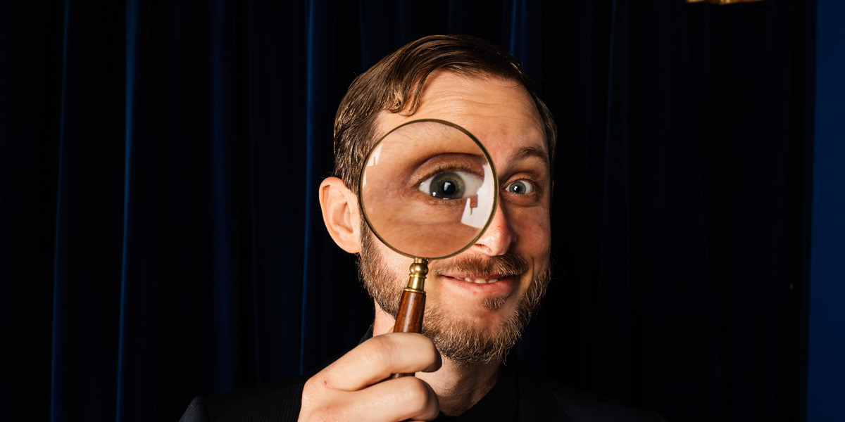 A magician holds a magnifying glass up to his eye, an enlarged image of his eye is visible in the magnifying glass.