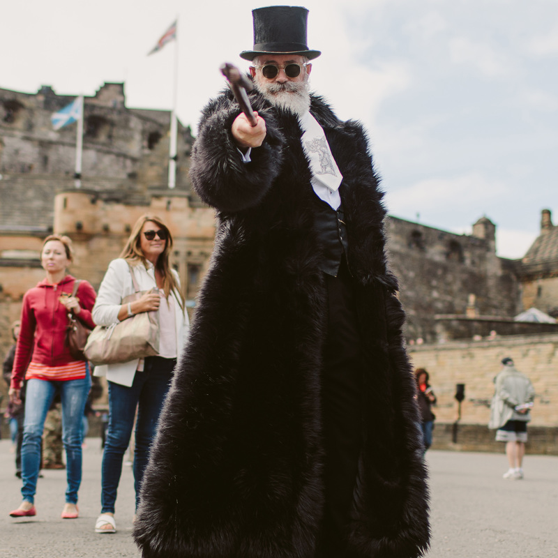 A bearded gentleman is pointing a walking cane toward the camera outside of Edinburgh castle, he is wearing a top hat and a large black fur coat and is carrying a case with Werewolves written on it. Members of the public are observing him.