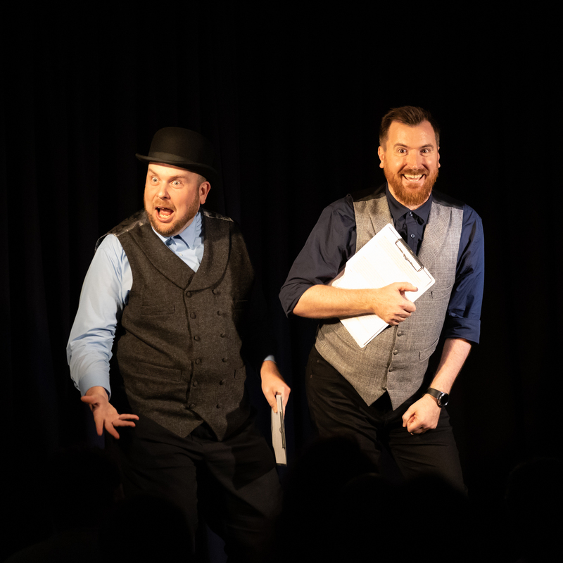 Two well dressed men are smiling happily on stage as they entertain a packed theatre.