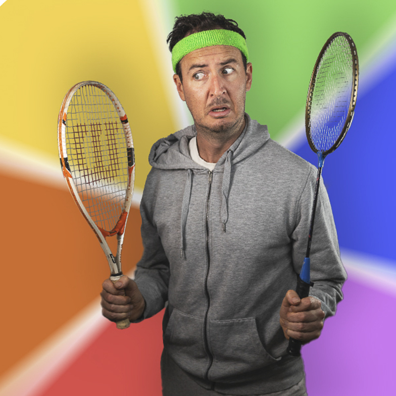 A tall man with dark hair in a gray tracksuit with a green headband looks puzzled at a tennis racquet in one hand and a squash racquet in the other. The background is the different coloured wheels of a Trivial Pursuit pie wheel from the board game