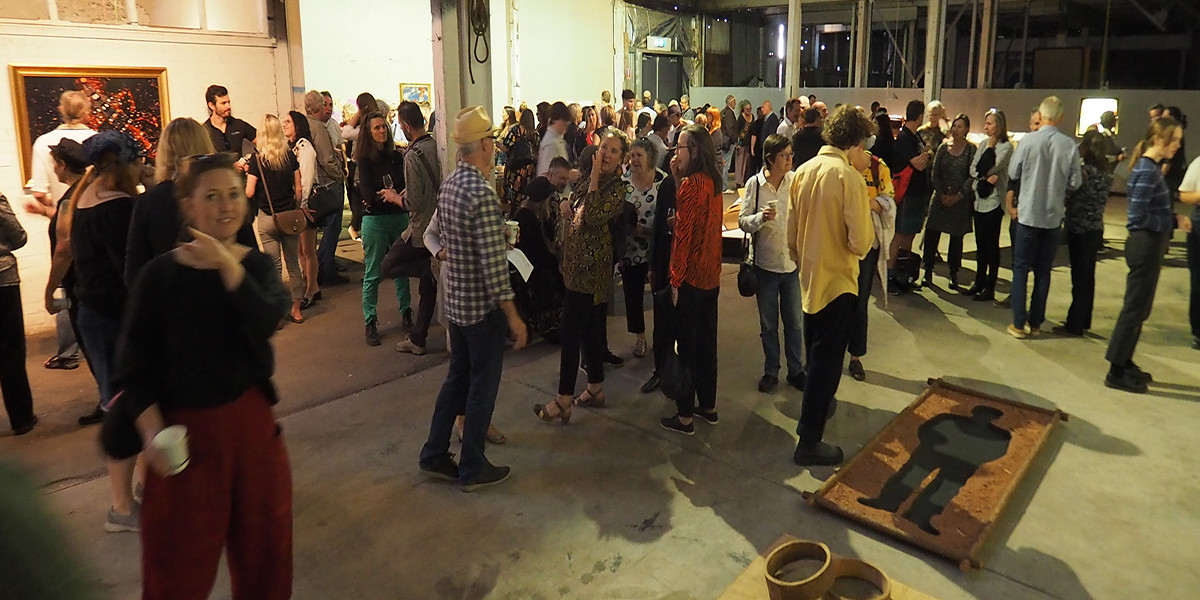 A photo of part of the crowd who came to the opening of "Vessel" in 2020. On the left can be seen a gold framed photograph by Narelle Autio titled "Goana Number 1, 2002" showing the part of a dead body of a goanna from a series of road kill images  called "Indifference". In the foreground to the right can be seen a part of Hossein Valamanesh's installation from 1985 titled "Sifting through, gold was found here", mixed media. The crowd are standing around inside this beautiful industrial space, chatting with each other.