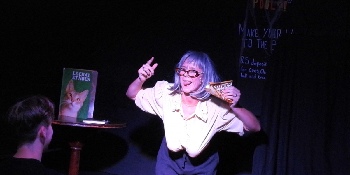 A young woman dressed as old lady in a grey wig, brown glasses and cream blouse, holds a packet of Werther's Original sweets in her left hand. her tongue is out and her right hand points to the packet. A green book with a cat on it sits on the table in the background. She is on stage, mid show.