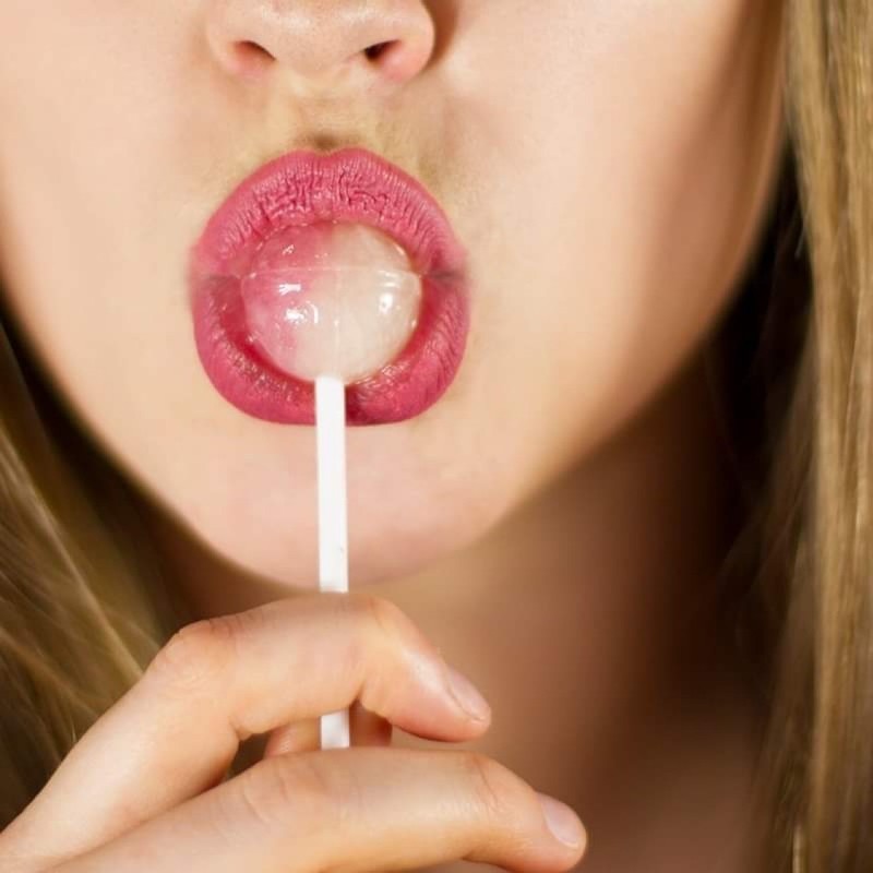 SUGAR - Female pink lips with a lollipop in focus, sandy blonde hair and her hand slightly blurry in the background.