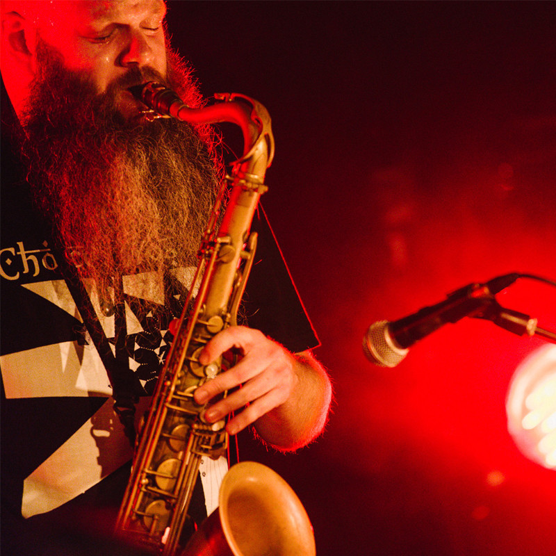 A man plays the saxophone with his eyes closed and head tilted, he is on the left hand side facing the right with a warm red light reflecting off his face and another red glowing light in the background to the right. He has a long brown and white bead and wears a black t-shirt with white graphics, there is a microphone positioned to the right of him in front of the saxophone.
