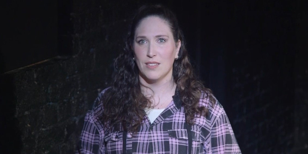 A pale-skinned woman with brown, curly hair and a purple checkered flannelette shirt with a concerned expression on her face