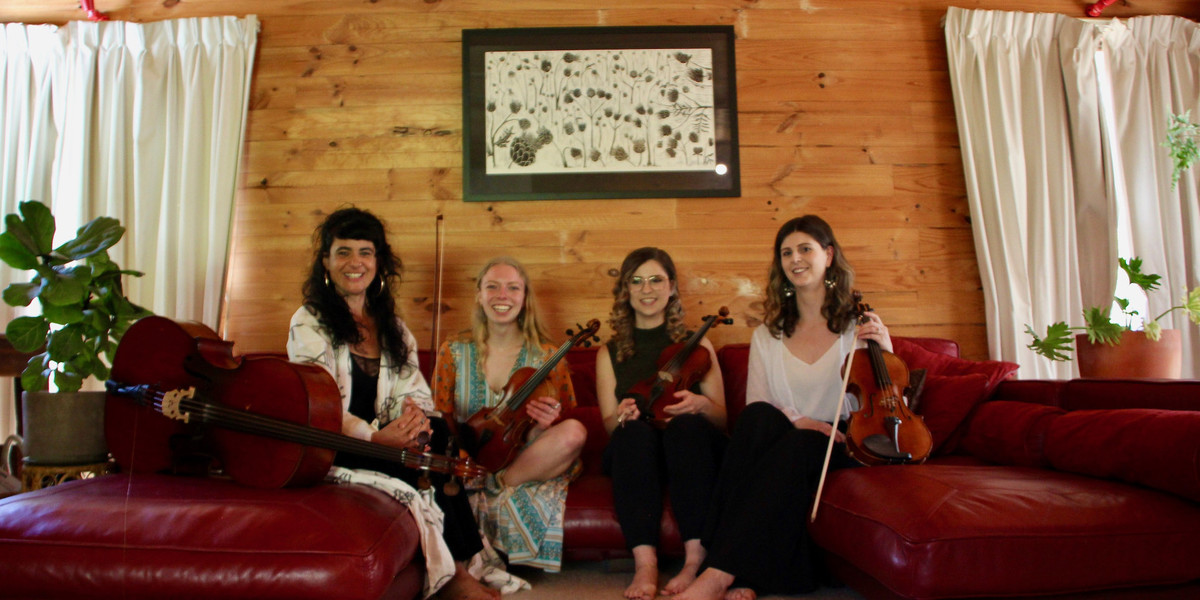 Four women sit on a red leather couch, on each side sits a green plant. All four women have their instrument in front of them; cello, two violins, and a viola. All smiling ear to ear.