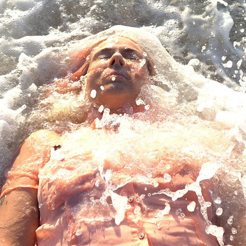 A photo of a person lying down while water crashes over them. They are wearing a light orange coloured shirt.