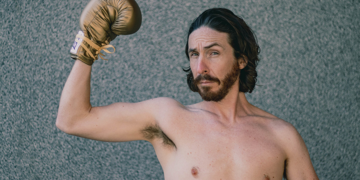 A bearded, shirtless man wears a golden boxing glove and flexes his right arm