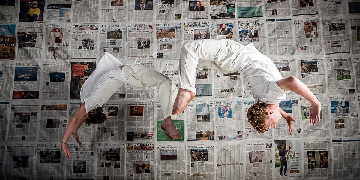 Two teenage male acrobats dressed in white are photographed in mid-air while performing a back-flip.