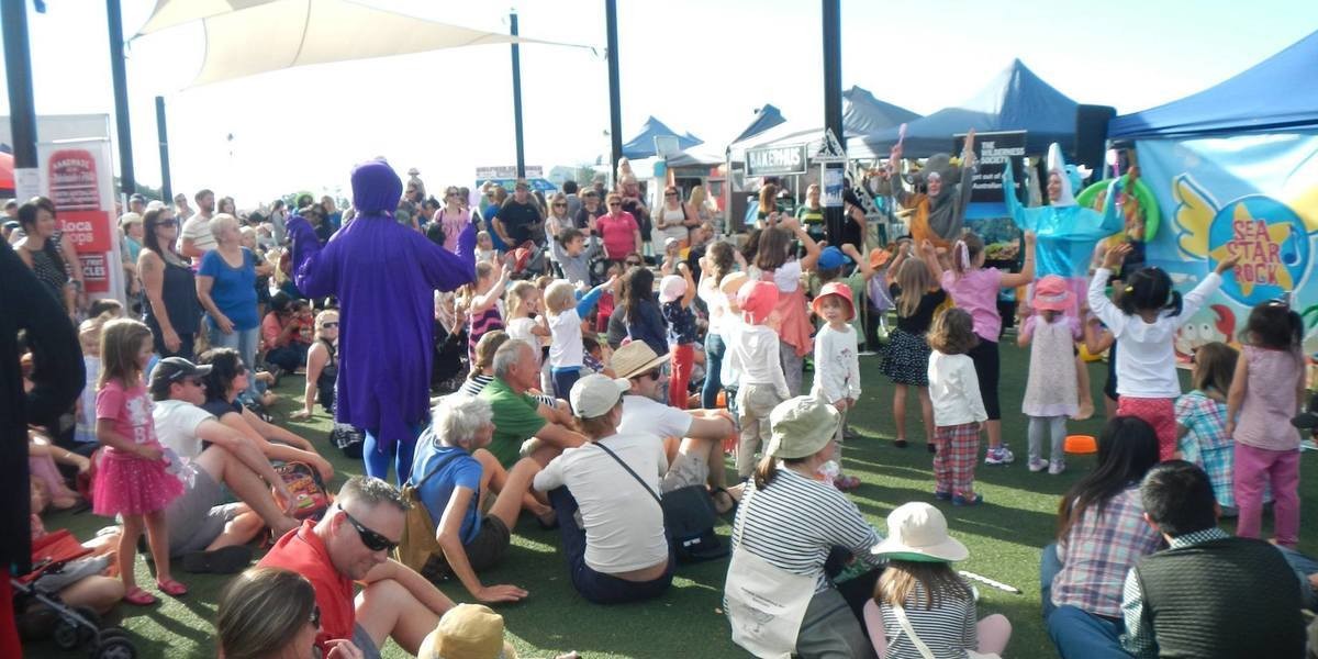 A SeaStar rock banner in the background with performers in front. One is dressed as a penguin and a dolphin. Kids and families are dancing outside to the music.