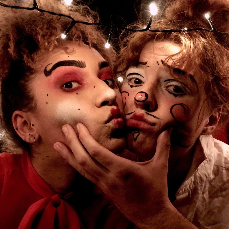 A photo of two people pouting their lips together and with one hand holding their faces together. Both people have intricate face makeup.