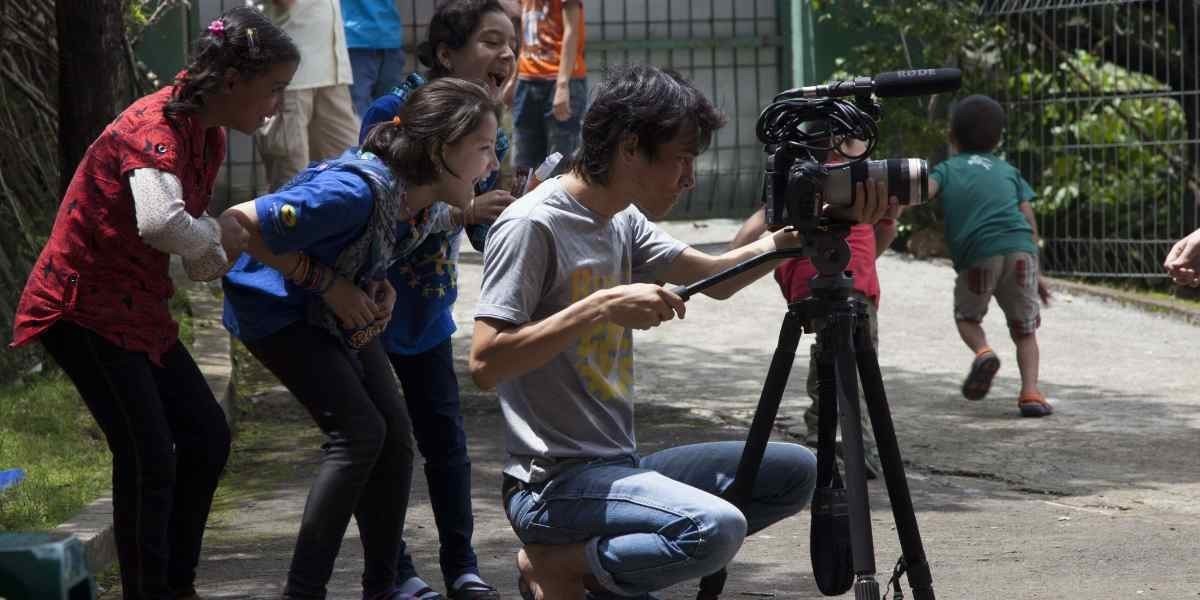 A young man films with a video camera while two young girls watch, laughing.