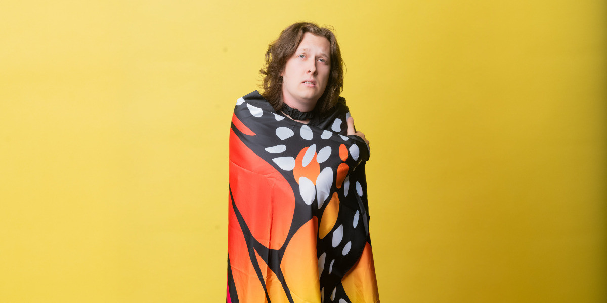 Jon wraps themselves in butterfly wings. They are in front of a yellow background.