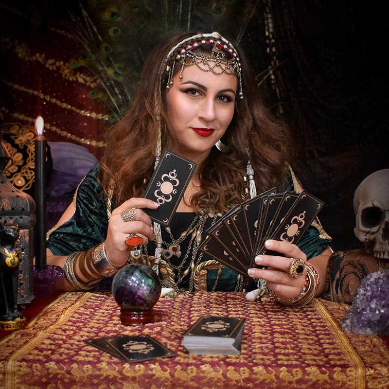 woman dressed as a gypsy, with jewellery and green velvet dress. Holding tarot cards, with a mysterious smile. On her table is a lit candle, tarot cards, a labradorite crystal ball, amethyst crystal, skull and a ornate red and gold cloth