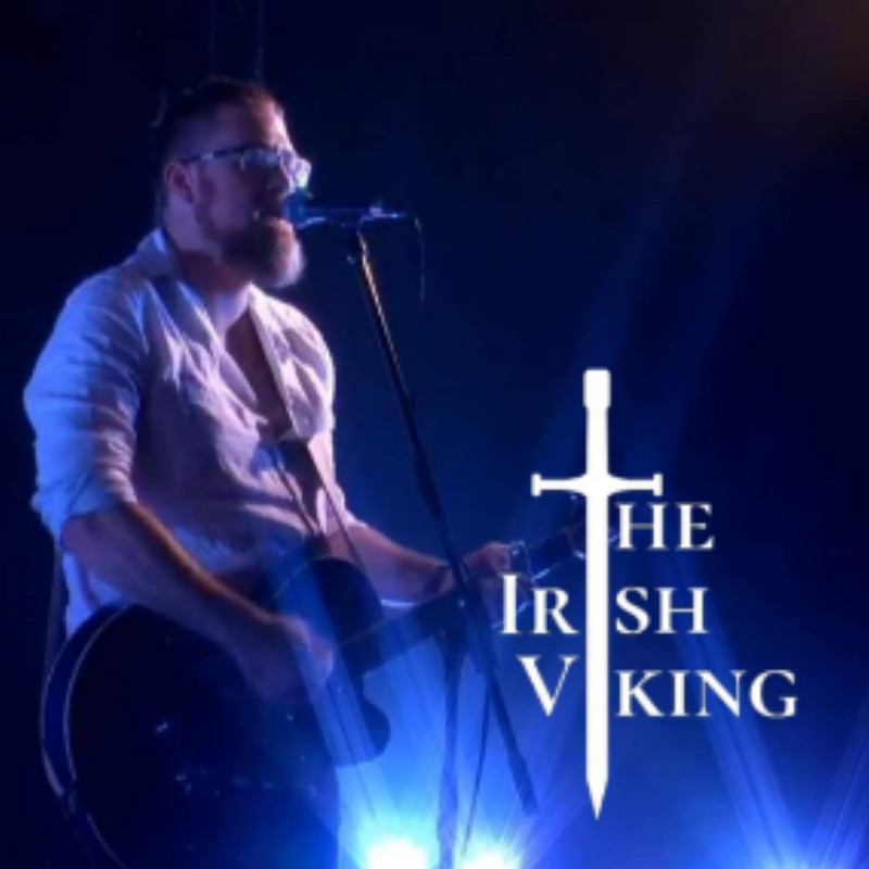 A Night in Valhalla - A photograph of a man performing on stage. He is singing into a microphone and playing guitar. He is wearing a light coloured shirt and glasses. The light from behind him is blue. On the photograph the text reads, ‘The Irish Viking’ in white font, with an image of a sword forming the ‘T’ ‘I’ and ‘I’ in each word.