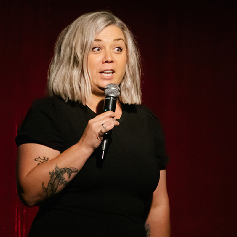 Image of Comedian, Nicky Wilkinson, live on stage. She is wearing a black top and is holding a microphone. She has an expressive face, as if mid-joke.