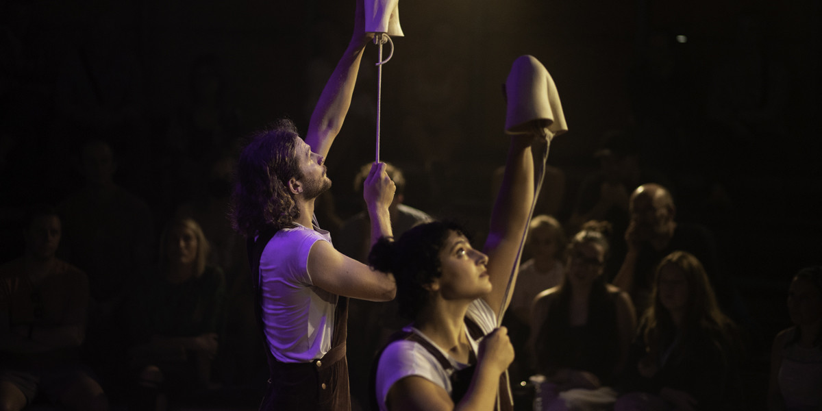 A dark image from the show, Both performers hold a ceramic elbow sculpture above their head.