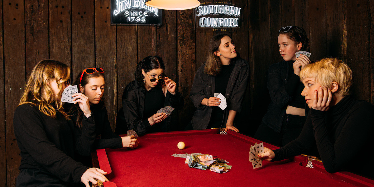 a group of 6 females sit around a red pool table, playing a game of cards, all of which are suspicious of each other and what they're next move is.