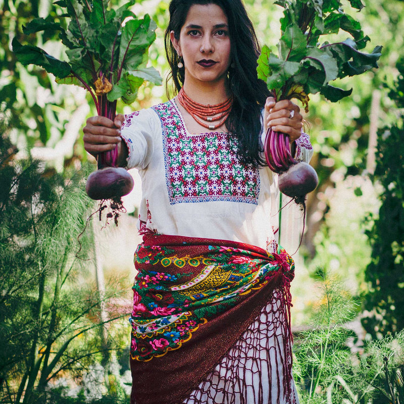 A performer with long dark skin, hair and eyes looks stoically at the camera while standing in a lush green garden. She is wearing ethnic clothing and is holding in her outstretched hands two large beetroots