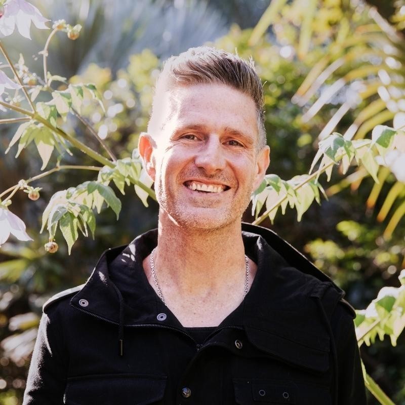 A mid shot of Wil Anderson standing outside in the sun in front of green bushes. He's smiling a wearing a back denim jacket.