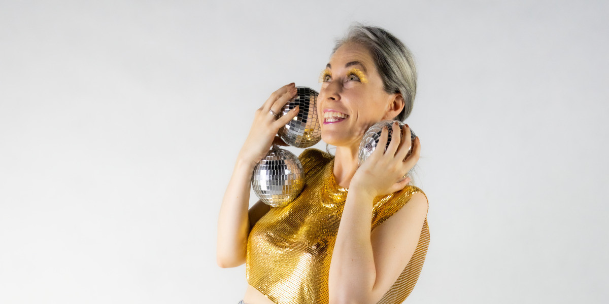 Laughing woman in gold shirt and gold lashes holds three mirror (disco) balls up to her face