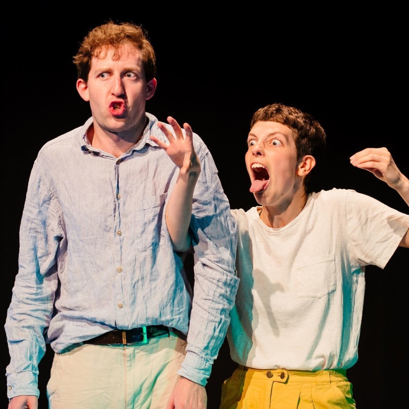 A photo of two people with animated expressions on their faces. The person on the left is wearing a light blue long sleeved shirt and cream coloured pants. The person on the right is wearing a white t-shirt and yellow pants.