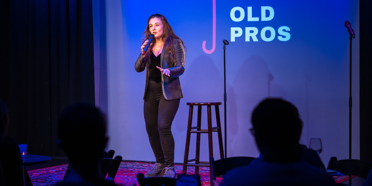 Kaytlin Bailey, mid-30s white cis-woman with brown long hair wearing a jeans and a leather jacket on stage holding a microphone next to a stool, with a blue background