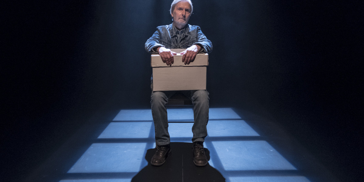 A man sits on a chair on a dark stage with a soft light over him as he holds a cardboard box.
