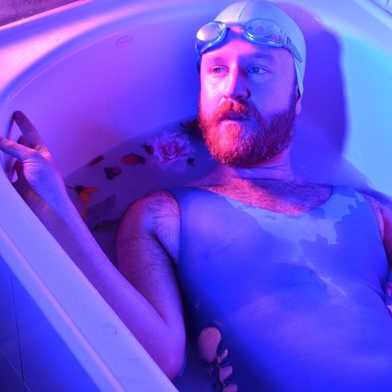 A photo of a man wearing a blue swimsuit sitting in a bathtub. He has a full beard and is wearing a blue swimming cap and googles on his head.