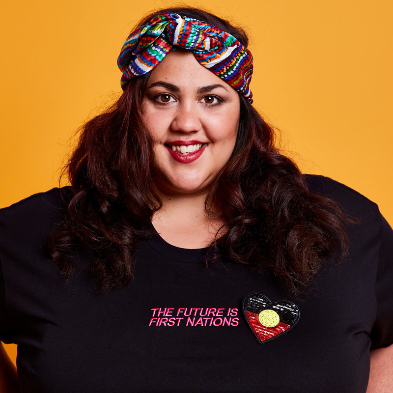 Steph Tisdell stands proudly, smiling, hands on hips wearing a black t-shirt that says "The Future is First Nations" with an Aboriginal Flag heart shaped pin.