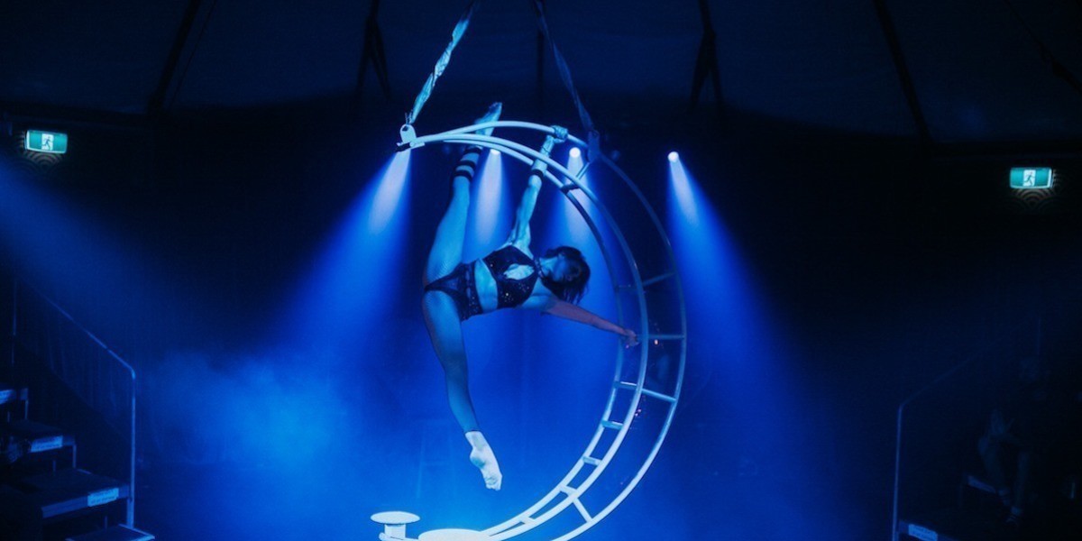 Aerial performer is holding her body in full splits across a giant metal crescent moon apparatus, which is suspended from the tent center stage and lit from behind.