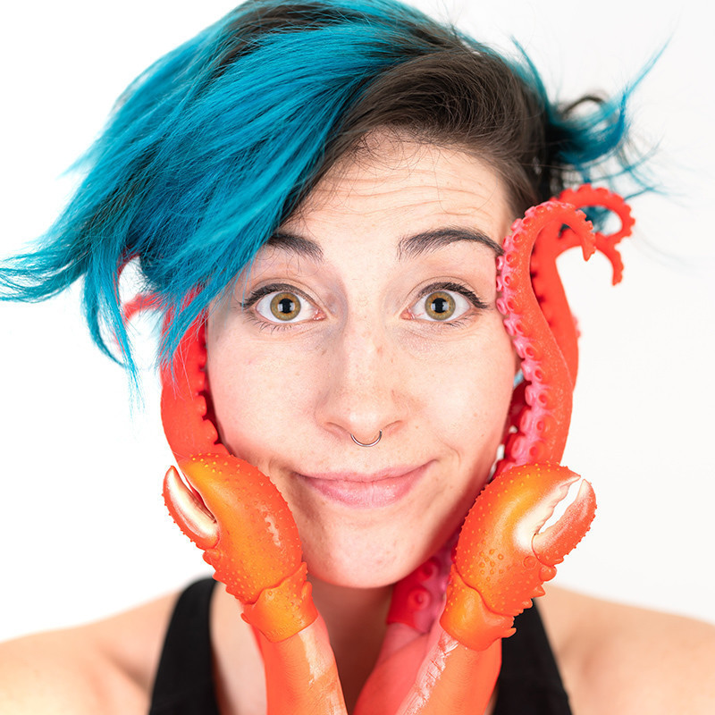 A photo of a person with blue hair and orange tentacle like fingers placed on the side of their face.