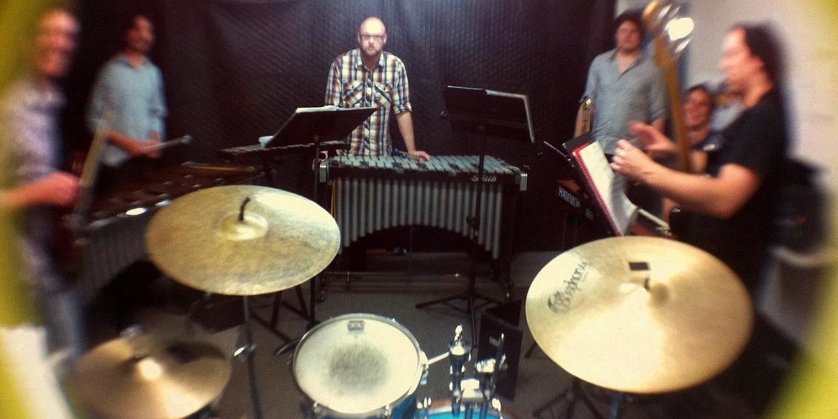 A photograph of five people standing around a drum kit.